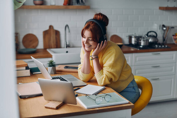 Радостная молодая женщина в наушниках смотрит на ноутбук, сидя за кухонным столом