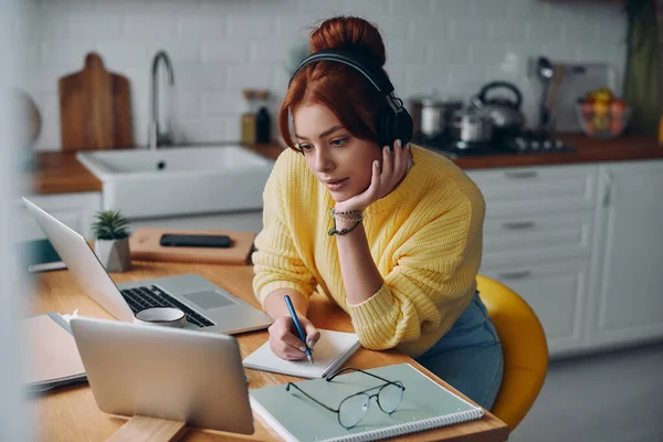 ヘッドフォンで美しい女性はノートを作り キッチンカウンターに座っている間にラップトップを見て — ストック写真