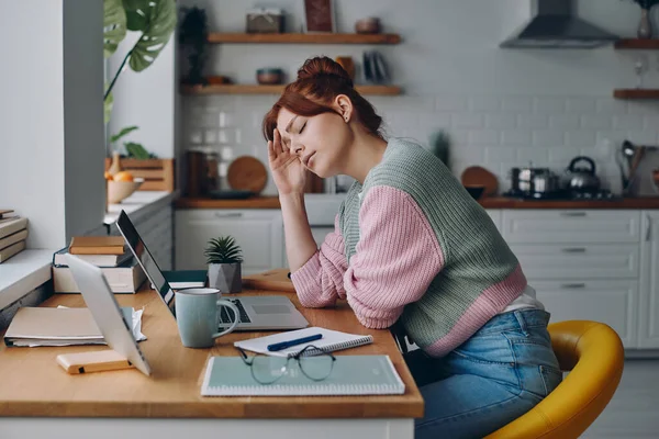 Frustrated young woman touching head while working from home at the domestic kitchen