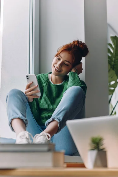 Hermosa Mujer Joven Mirando Teléfono Inteligente Sonriendo Mientras Sienta Alféizar — Foto de Stock