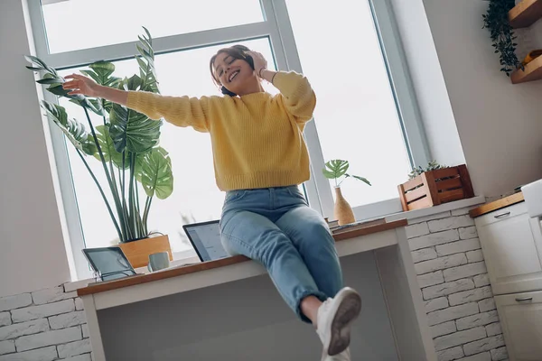 Excited young woman in headphones gesturing while sitting on the kitchen counter at home