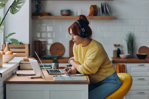 stock image Confident woman in headphones making notes and looking at laptop while sitting at the kitchen counter