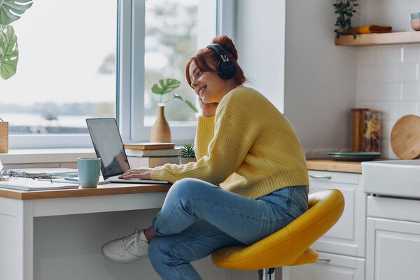 Красивая молодая женщина в наушниках с помощью ноутбука и улыбаясь, сидя за кухонным столом