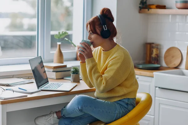 Vacker Kvinna Hörlurar Dricker Kaffe Och Tittar Laptop Medan Sitter — Stockfoto