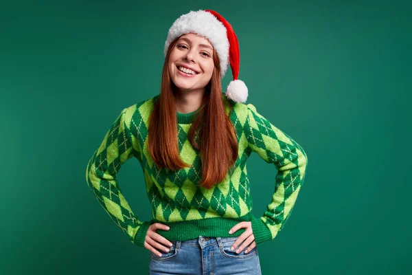 美しい若いです女性でクリスマス帽子保持手でヒップと笑顔に対して緑の背景 — ストック写真
