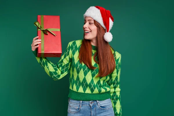 戴着圣诞礼帽的年轻漂亮女人看着礼品盒 在绿色的背景下微笑着 — 图库照片