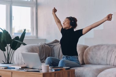 Evde dizüstü bilgisayarın yanında otururken kollarını kaldıran kulaklıklı mutlu genç bir kadın.