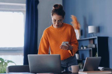 Ofiste çalışırken cep telefonu tutan ve dizüstü bilgisayar kullanan kendinden emin genç bir kadın.