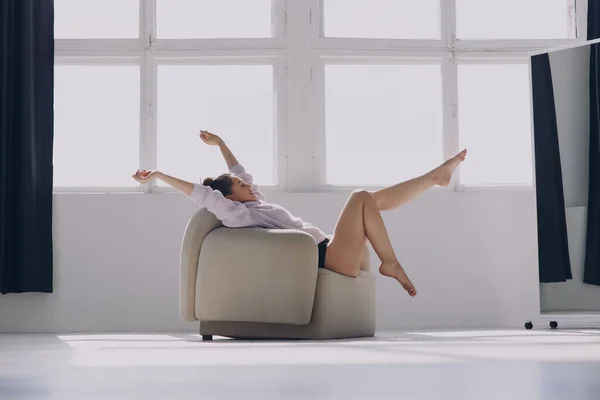 窓の前の快適な椅子でリラックスしながら腕を上げ続ける美しい若い女性 — ストック写真