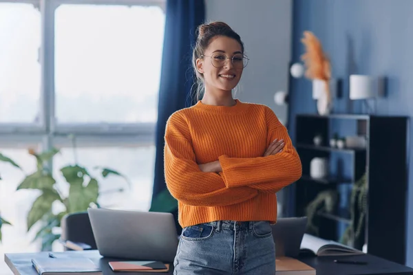 Confident woman keeping arms crosses and smiling while standing near her working place in office