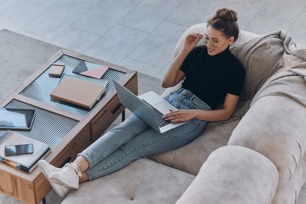 Top view of beautiful young woman using laptop while sitting on the comfortable couch at home