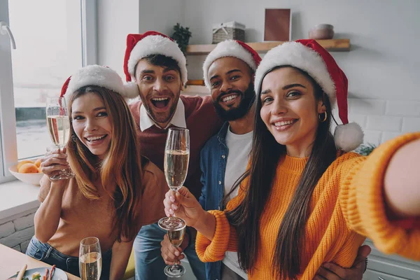 クリスマスの帽子の美しい人々は自宅で自画自賛しながらシャンパンフルートを保持 — ストック写真