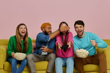 Bir grup neşeli genç, pembe arka planda kanepede otururken video oyunu oynuyor.