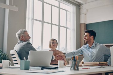 İş adamları el sıkışıyor ve ofis masasında birlikte otururken mutlu görünüyorlar.