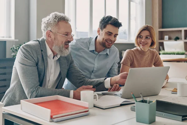 Τρεις Σίγουροι Επιχειρηματίες Κοιτάζοντας Φορητό Υπολογιστή Και Χαμογελώντας Ενώ Εργάζονται — Φωτογραφία Αρχείου