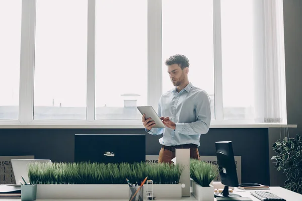 オフィスの窓の近くに立っている間 デジタルタブレットを使用して若いビジネスマンを自信を持って — ストック写真
