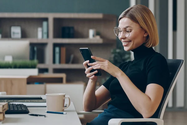オフィスの彼女の職場に座っている間 スマートフォンを使用して幸せな若いビジネスマンの女性 — ストック写真