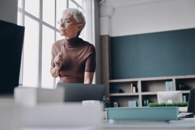 Ofisteki iş yerinin yanında duran kıdemli iş kadını düşünceli görünüyor.