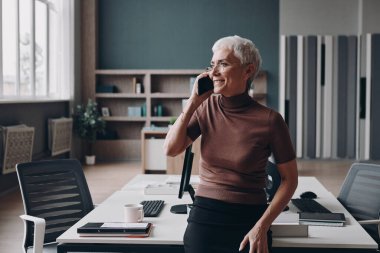 Ofis masasına yaslanırken cep telefonuyla konuşan kıdemli iş kadını.