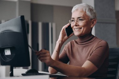Güzel kıdemli iş kadını ofisinde otururken akıllı telefon kullanıyor.