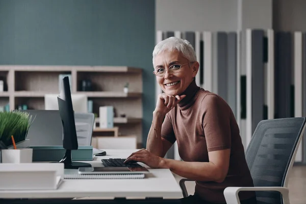 Starsza Bizneswoman Patrzy Obiektyw Uśmiecha Się Siedząc Biurze Miejscu Pracy — Zdjęcie stockowe