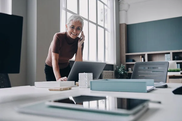 オフィスのノートパソコンで仕事をしながら携帯電話で話している幸せな先輩ビジネスマンの女性 — ストック写真