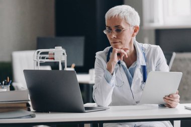 Tıp bürosundaki masada otururken teknolojilerini kullanan kendine güvenen bir kadın doktor.