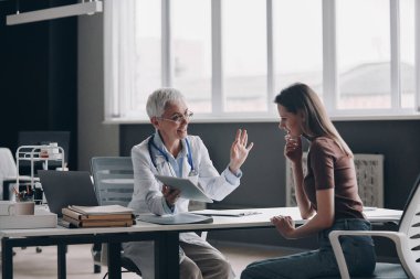 Kendine güvenen kadın doktor hastayla konuşuyor ve ofiste birlikte otururken gülümsüyor.