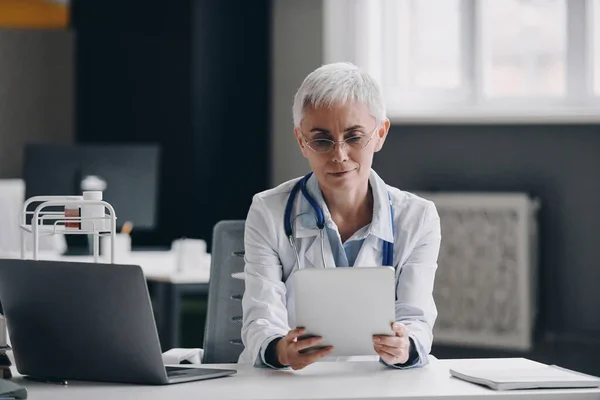 Médica Confiante Olhando Para Tablet Digital Enquanto Sentada Mesa Consultório — Fotografia de Stock
