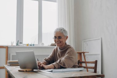 Kıdemli iş kadını dijital tablet kullanıyor ve çalıştığı yerde otururken gülümsüyor.