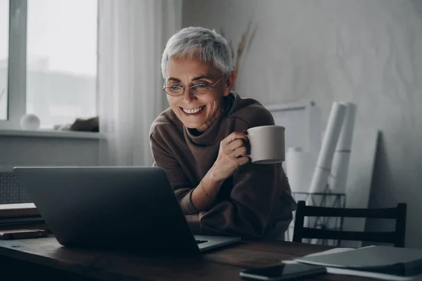Ευτυχισμένη Ηλικιωμένη Γυναίκα Που Χρησιμοποιεί Φορητό Υπολογιστή Και Απολαμβάνει Ζεστό — Φωτογραφία Αρχείου