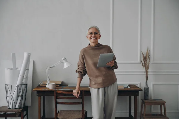 一位快乐的资深女商人站在自己的工作岗位旁 手里拿着数码平板电脑 — 图库照片