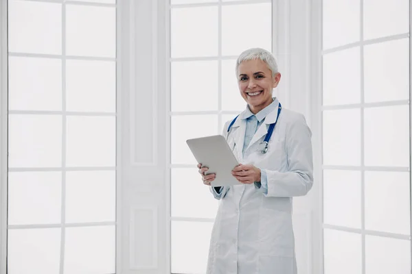 ウィンドウに対して立っている間にデジタルタブレットと笑顔を保持している自信の女性医師 — ストック写真