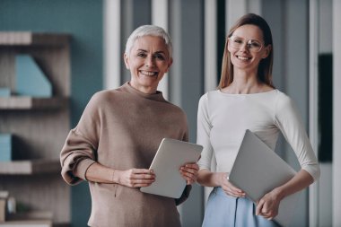 İki kendine güvenen iş kadını ellerinde aletlerle ofiste yan yana dururken gülümsüyorlar.