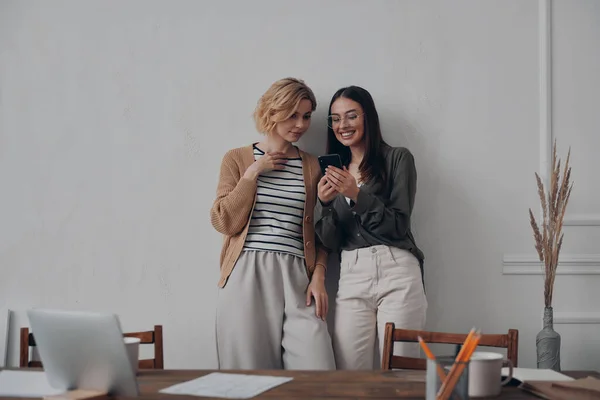創造的な職場の近くに立っている間 スマートフォンを使用して2人の美しい若い女性 — ストック写真