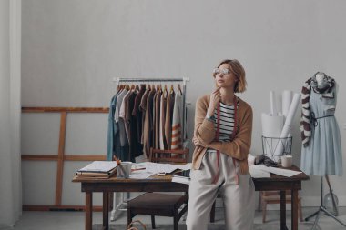 Çekici bayan moda tasarımcısı atölyedeki masaya yaslanırken düşünceli görünüyor.