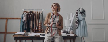 Güzel bayan moda tasarımcısı atölyede masaya yaslanırken düşünceli görünüyor.
