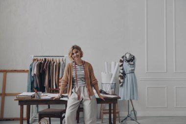 Kadın moda tasarımcısı kameraya bakıyor ve işyerinin yanında dururken gülümsüyor.