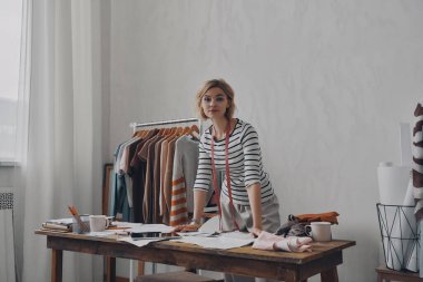 Genç bayan moda tasarımcısı iş yerinin yanında dururken kameraya bakıyor.