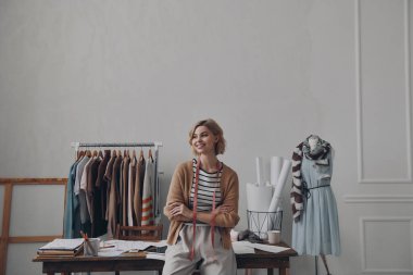 Atölyede masaya yaslanırken gülümseyen kendine güvenen kadın moda tasarımcısı.