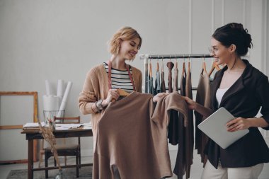 İki kadın moda tasarımcısı atölyede çalışırken kıyafetleri tartışıyor.