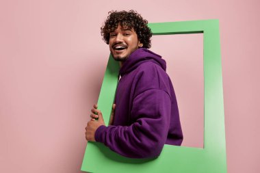 Mutlu Hintli adam pembe arka planda dikilirken çerçeveden dışarı bakıyor.