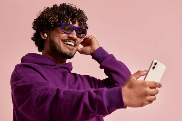 快乐的印度人一边调整耳机 一边在粉色背景下使用智能手机 — 图库照片