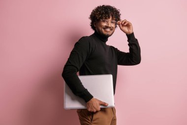 Dizüstü bilgisayar taşıyan ve pembe arka planda gözlük takan mutlu Hintli adam.