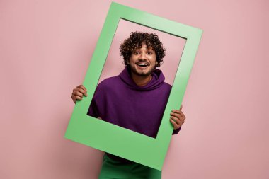 Mutlu Hintli adam pembe arka planda dikilirken çerçeveden dışarı bakıyor.