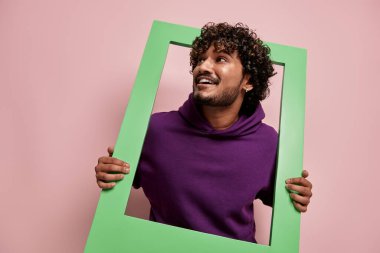 Heyecanlı Hintli adam pembe arka planda dikilirken çerçeveden dışarı bakıyor.