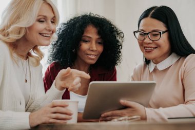 Masada birlikte otururken dijital tablete bakan üç mutlu olgun kadın.