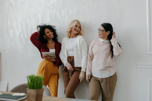 オフィスの壁にもたれている間 3人の自信に満ちた成熟した女性が話して笑顔 — ストック写真