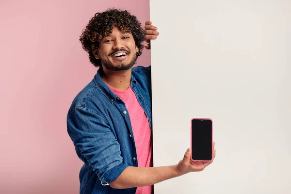 快乐的印度人一边展示智能手机 一边靠在粉色背景的复制空间上 — 图库照片