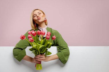 Güzel genç bir kadın elinde lalelerle pembe arka planda gülümsüyor.